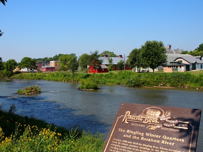 [Looking across a somewhat shallow river at brick buildings on the other side. A plaque describing the scene is in the foreground in the lower right.]